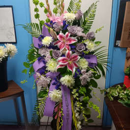 Tall floral spray with pink lilies, lavender roses, and purple ribbon in a black vase