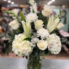 White floral arrangement in a clear glass vase with roses, lilies, and hydrangeas