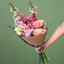 Handheld bouquet of pink, peach, and lavender flowers wrapped in brown paper