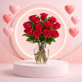 Red roses in a clear glass vase with pink heart balloons around them.