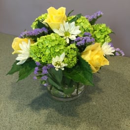 Yellow roses and white daisies in a glass bowl with green hydrangeas and purple filler flowers