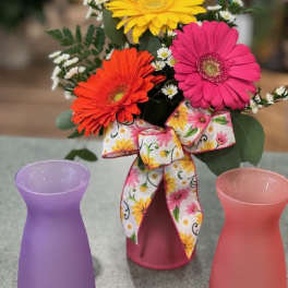 Bright gerbera daisies in a frosted pink vase with floral bow, flanked by two empty frosted vases.