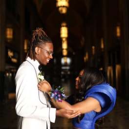Couple in formal attire exchanging a boutonniere in a dim hallway