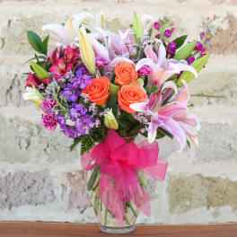 Bouquet of lilies, roses, and purple flowers in a glass vase with a pink ribbon