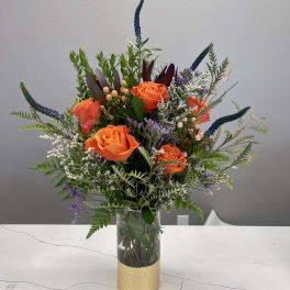 Orange roses in a clear glass vase with mixed greenery and purple accents