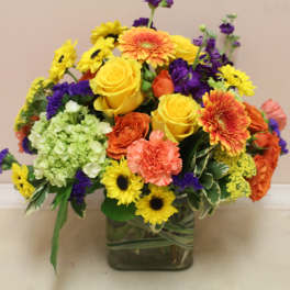 Mixed bouquet of yellow, orange, purple, and green flowers in a square glass vase