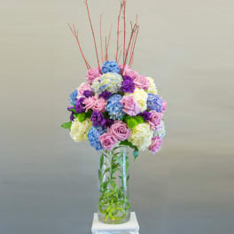 Tall bouquet of pink roses, blue hydrangeas, and purple flowers in a glass vase