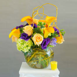 Bouquet of yellow calla lilies, roses, and purple blooms in a glass vase