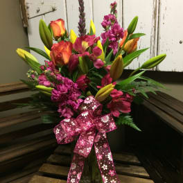 Mixed bouquet of lilies, roses, and purple blooms in a glass vase with a pink ribbon