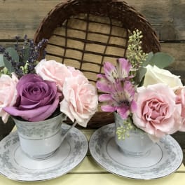 Two small floral arrangements in teacups on saucers with pink and purple roses