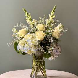 Arrangement of pale yellow roses, white hydrangeas, and snapdragons in a clear glass vase.