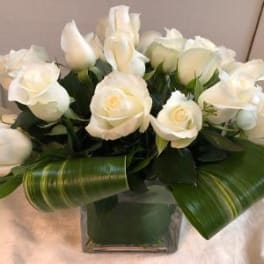 White roses arranged in a square glass vase with broad green leaves