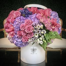 Pink roses and orchids arranged with lavender hydrangeas in a white hatbox