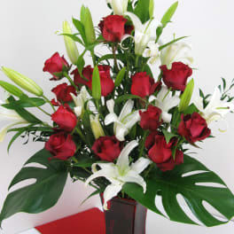Tall arrangement of red roses and white lilies with large tropical leaves in a red glass vase.