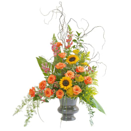Tall arrangement of orange roses, sunflowers, and pink snapdragons in a dark pedestal vase