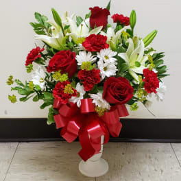 Red roses, white lilies, and daisies arranged in a white vase with a red ribbon.
