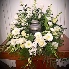 White funeral floral arrangement around a gray urn on a wooden table