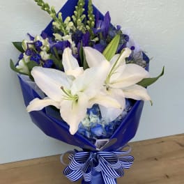 Bouquet of white lilies and blue flowers wrapped in blue paper with a striped ribbon.