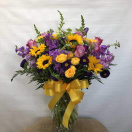Bouquet of sunflowers, purple flowers, and pink roses in a glass vase with a yellow ribbon.