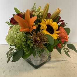 Mixed bouquet with orange lilies, a sunflower, and roses in a square glass vase