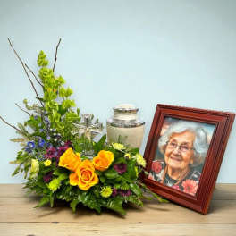 Yellow roses and mixed flowers arranged beside a framed portrait and urn.