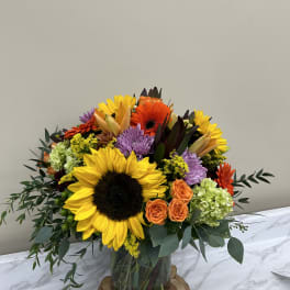 Mixed bouquet with sunflowers, orange roses, and purple blooms in a glass vase