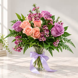 Pink and lavender roses arranged in a glass vase with a purple ribbon