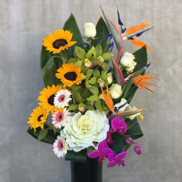 Tropical arrangement with sunflowers, white roses, orchids, and bird-of-paradise in a black vase.