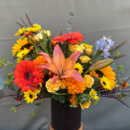 Mixed bouquet with a pink lily, gerbera daisies, sunflowers, and yellow roses in a dark vase
