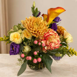 Mixed bouquet with yellow roses, orange calla lily, and purple blooms in a glass vase