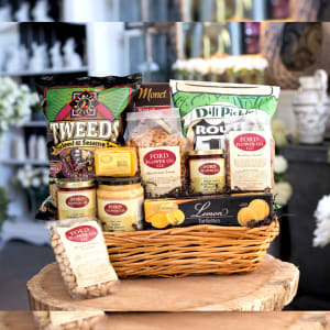 Wicker gift basket filled with assorted packaged snacks and gourmet treats on a wooden surface.