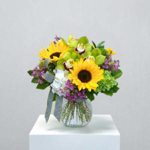 Bright arrangement of yellow sunflowers, green orchids, and mixed blooms in a clear glass vase with checked ribbon.