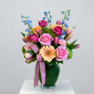 Colorful bouquet of pink roses, tulips, gerbera daisies, and blue delphinium in a green vase with ribbon.