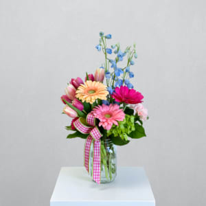 Mixed pink gerbera daisies, tulips, and blue delphinium in a mason jar with gingham ribbon