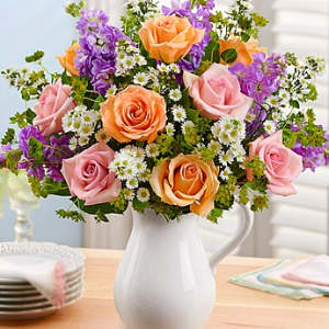 Pink and peach roses with purple flowers in a white pitcher vase