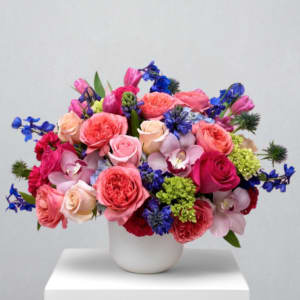 Low arrangement of pink roses, blue delphinium, and mixed flowers in a white round vase