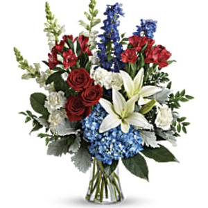 Tall red, white, and blue flower arrangement with roses, lilies, hydrangea, and delphinium in a clear glass vase