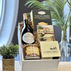 Gourmet gift crate with wine, crackers, peanuts, chocolates, and snack mixes on a wooden surface