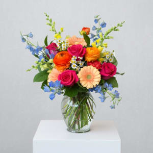Bright mixed bouquet of pink roses, orange blooms and blue flowers in a clear glass vase.