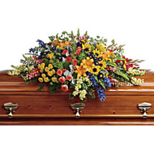 Bright mixed flower casket spray in yellow, orange, blue, and pink on a polished wooden casket