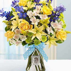 Bouquet of yellow roses, lilies, and blue flowers in a glass vase with a baby photo frame