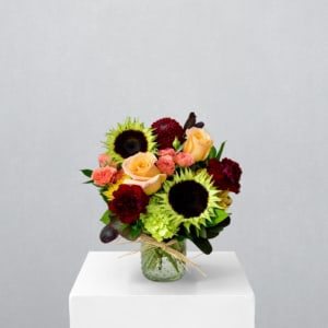 Bouquet of sunflowers, peach roses, and burgundy blooms in a glass vase