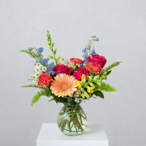 Mixed bouquet of bright pink, peach, yellow, and blue flowers in a clear glass vase.