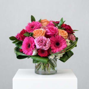 Low round arrangement of pink gerbera daisies and roses with peach accents in a clear glass vase.