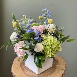 Mixed bouquet in a white square vase with pink roses, green hydrangea, and blue flowers