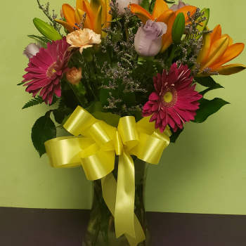 Mixed bouquet of pink gerbera daisies, orange lilies, and roses in a glass vase with a yellow ribbon