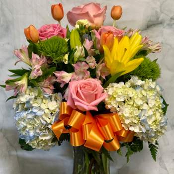 Tall mixed bouquet of pink roses, yellow lilies, tulips and hydrangeas in a clear vase with an orange bow.