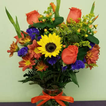 Mixed bouquet of orange roses, sunflowers, and purple flowers in a glass vase