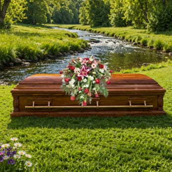 Pink and white casket spray with lilies and roses on a wooden casket in a grassy riverside setting