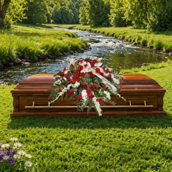 Red and white funeral spray on a wooden casket beside a river in a green meadow.